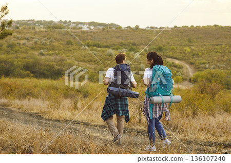 Hikers couple with backpacks hiking on path through field and meadow, back view Hikers couple with backpacks hiking on path through field and meadow, back view 136107240