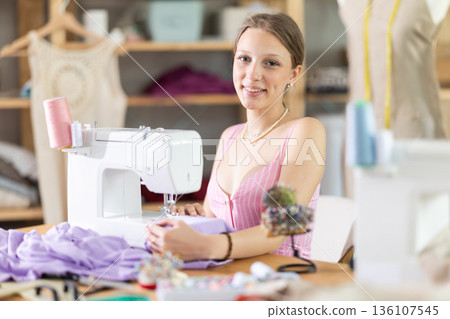Positive young female dressmaker working with sewing-machine in tailor's workshop Positive young female dressmaker working with sewing-machine in tailor's workshop 136107545