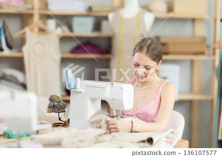 Young seamstress sews a dress on a machine 136107577