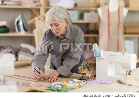 Mature female dressmaker drawing on paper in sewing workshop Mature female dressmaker drawing on paper in sewing workshop 136107592