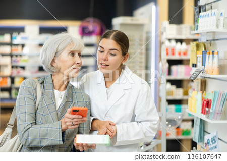 Mature female buyer together with pharmacist scans QR code on medicine label in pharmacy Mature female buyer together with pharmacist scans QR code on medicine label in pharmacy 136107647