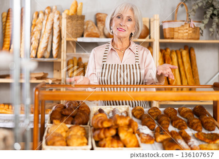 Portrait of elderly woman baker standing behind display case with fresh croissants. Offering to buy fresh baked goods Portrait of elderly woman baker standing behind display case with fresh croissants. Offering to buy fresh baked goods 136107657