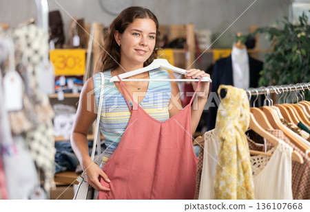 Positive young woman choosing something for summer in clothing store 136107668