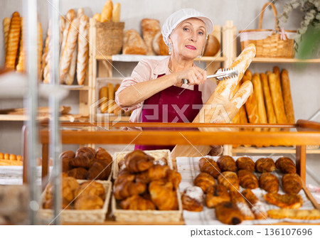 Mature woman bakery employee offering bread and different baguettes for sale Mature woman bakery employee offering bread and different baguettes for sale 136107696