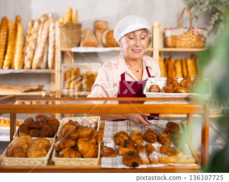 Senior woman employee puts croissants in window, arranges display of goods at bakery. 136107725