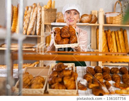 Positive elderly female seller with tongs in his hands offers to buy fresh croissants Positive elderly female seller with tongs in his hands offers to buy fresh croissants 136107756