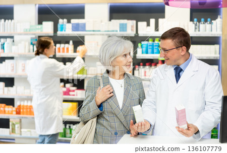 Middle-aged male pharmacist consulting old woman costumer about care product in box 136107779