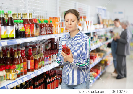 Woman customer is thinking about buying spicy and unusual red sriracha sauce. Woman customer is thinking about buying spicy and unusual red sriracha sauce. 136107791