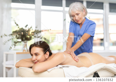 Blurred mature woman actively rubs lower back of young woman during general massage procedure. Blurred mature woman actively rubs lower back of young woman during general massage procedure. 136107832