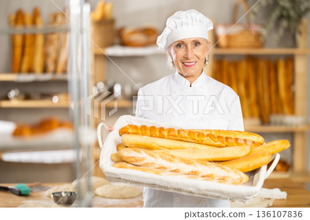 Positive elderly woman bakery employee offering bread and different baguettes for sale Positive elderly woman bakery employee offering bread and different baguettes for sale 136107836