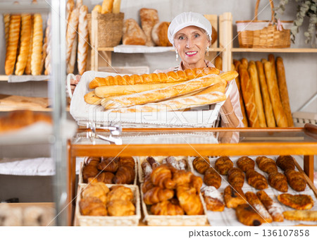 Senior female baker worker sells baguette, shows many products Senior female baker worker sells baguette, shows many products 136107858