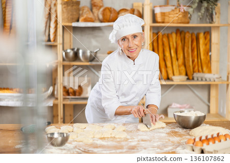Experienced elderly woman baker using knife to cut raw dough to make croissants Experienced elderly woman baker using knife to cut raw dough to make croissants 136107862