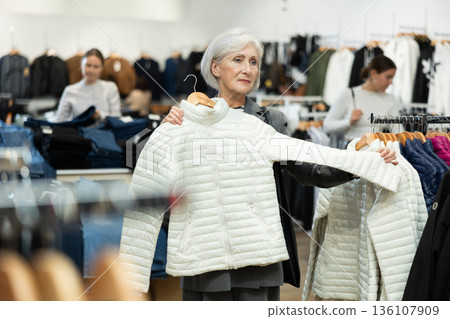 Elderly woman choosing white winter down jacket in clothing shop with large assortment Elderly woman choosing white winter down jacket in clothing shop with large assortment 136107909