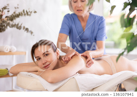 Blurred mature woman actively rubs shoulder of young woman during general massage procedure. 136107941