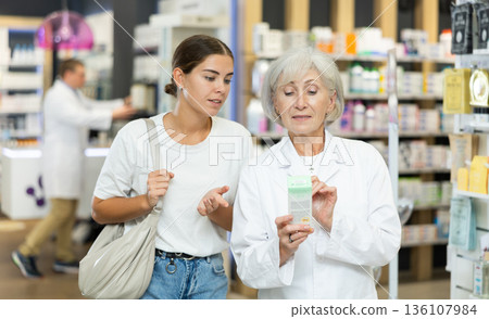 Old female pharmacist consulting young girl about care product in box 136107984