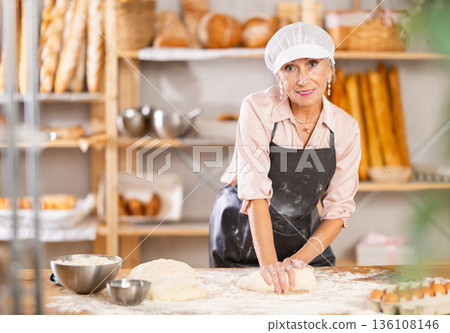 Senior woman works in bakery as baker, kneads dough, works with flour. 136108146