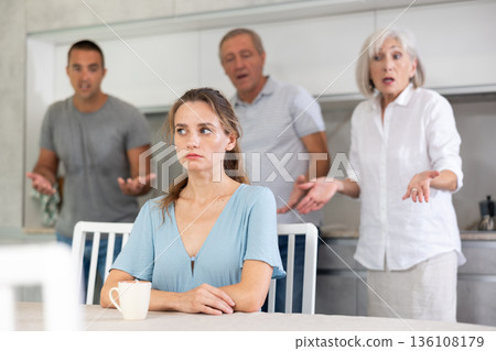 Domestic quarrel - offended young woman sits at table while parents and young husband yell at her 136108179