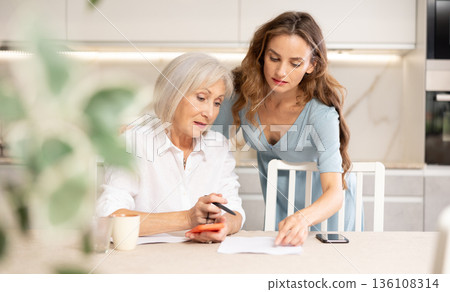 Mother and daughter sitting at table and calculating finances. Daughter using smartphone for online payment 136108314