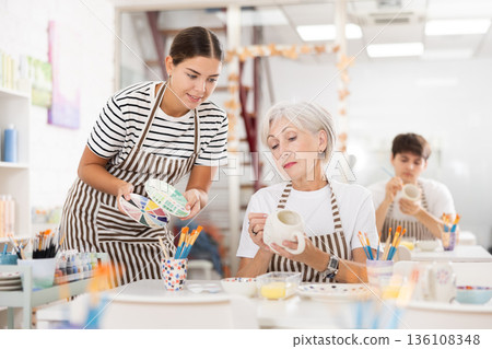 Young girl assisting senior woman painting ceramic mug in pottery class Young girl assisting senior woman painting ceramic mug in pottery class 136108348