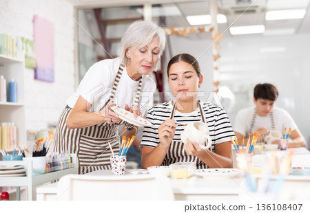 Senior woman assisting young girl painting ceramic mug in pottery class 136108407