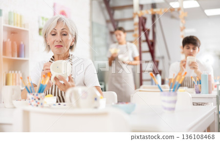 Talented senior female artisan applying design to ceramic mug in pottery studio Talented senior female artisan applying design to ceramic mug in pottery studio 136108468