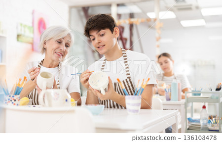 Cheerful guy talking with female colleague while painting mugs in pottery workshop 136108478