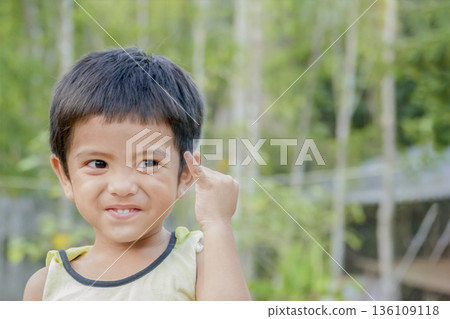Close up of smiling Asian little cute boy show pinkie finger or reconcile gesture concept and looking at camera outdoor over green nature background 136109118