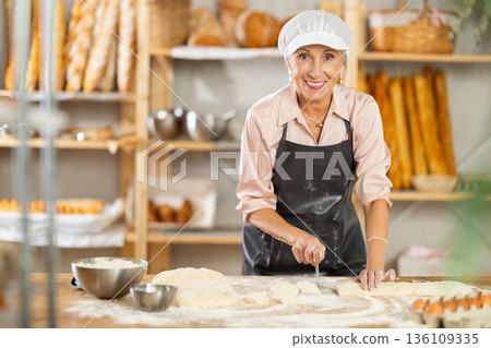 Professional elderly woman baker cutting raw dough with knife to make croissants Professional elderly woman baker cutting raw dough with knife to make croissants 136109335
