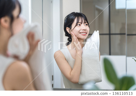 一位年輕的亞洲女性在鏡子前洗完臉後用毛巾擦乾臉（護膚，臉部護理） 136110513