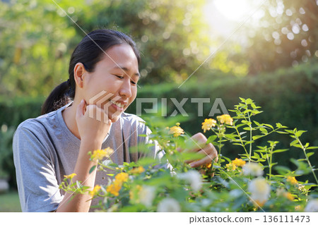 Close-up of a happy Asian woman smiling and enjoying purple flowers in her green garden. Beautiful female portrait in sunny backyard park. Concept of nature lovers, wellness, and outdoor lifestyle. 136111473