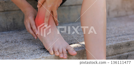 Close-up of woman hand holding painful injured ankle with red inflamed spot and bruising. Medical concept of joint pain, sprain, ligament injury, bone ache, or arthritis in outdoor background. Close-up of woman hand holding painful injured ankle with red inflamed spot and bruising. Medical concept of joint pain, sprain, ligament injury, bone ache, or arthritis in outdoor background. 136111477