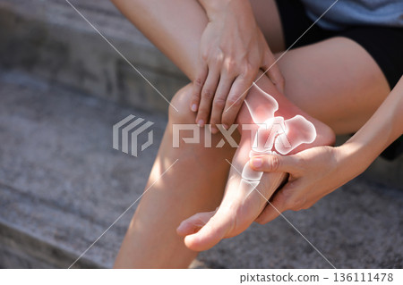 Close-up of hand holding painful injured ankle with red inflamed spot and X-ray bone skeleton joint overlay. Medical concept of fracture, sprain, arthritis, or ligament injury in outdoor background. 136111478