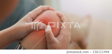 Close-up of child hand holding painful injured knee with red inflamed spot and X-ray bone skeleton joint overlay. Medical concept of pediatric fracture, arthritis, or muscle ache in indoor background. Close-up of child hand holding painful injured knee with red inflamed spot and X-ray bone skeleton joint overlay. Medical concept of pediatric fracture, arthritis, or muscle ache in indoor background. 136111494