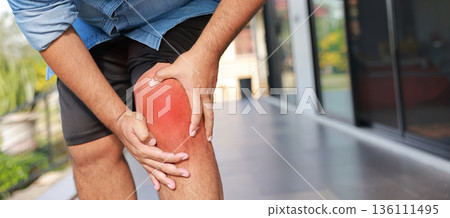 Close-up of hand holding painful injured knee with red inflamed spot and bruising. Medical concept of joint pain, arthritis, bone injury, muscle ache, or sprain in outdoor sunlight background. 136111495