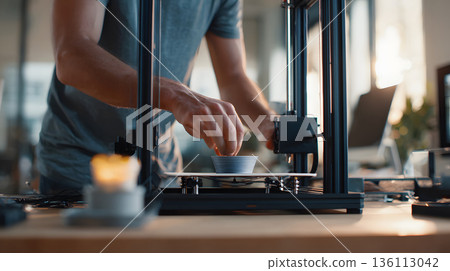 A person crafting a product using a 3D printer in a well-lit workspace. 136113042
