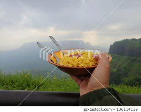 Hand holding a bowl of Indian snack with spoons, overlooking green valleys and cliffs in Matheran hill station, Maharashtra. 136114119