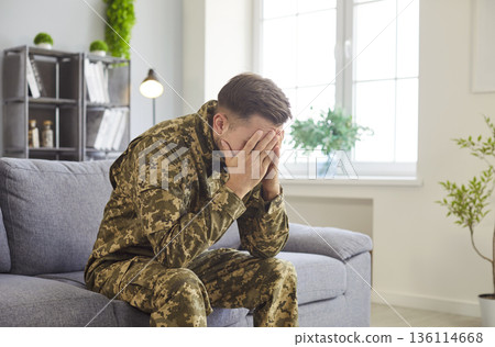 Young man in military camouflage uniform sits on couch expressing deep sadness or stress Young man in military camouflage uniform sits on couch expressing deep sadness or stress 136114668