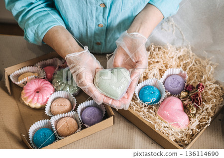 Hands hold heart shaped bath bomb over boxes filled with colorful handmade soaps and shredded paper. personalized beauty gift, diy spa kit, bespoke apothecary, aesthetic gift packaging. Hands hold heart shaped bath bomb over boxes filled with colorful handmade soaps and shredded paper. personalized beauty gift, diy spa kit, bespoke apothecary, aesthetic gift packaging. 136114690