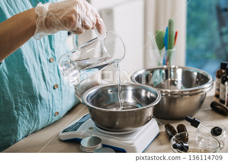 Person pours clear liquid from glass pitcher into metal bowl on digital scale. Cosmetic emulsion physics, oil and water mixing aesthetic, skincare manufacturing science, professional cream base making 136114709