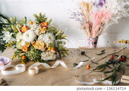 Flower bouquet featuring roses and chrysanthemums sits on brown paper near ribbon rolls. Sustainable floristry, plastic free gift wrapping, biodegradable materials, slow flower movement. Flower bouquet featuring roses and chrysanthemums sits on brown paper near ribbon rolls. Sustainable floristry, plastic free gift wrapping, biodegradable materials, slow flower movement. 136114714