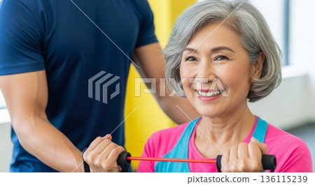 Senior asian woman doing resistance band exercise with fitness trainer guidance at gym physical therapy session 136115239