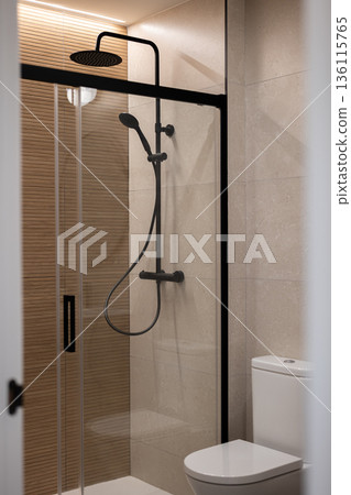 Modern bathroom with a glass shower enclosure, black fixtures, and beige tiles. A white toilet is visible in the corner. 136115765
