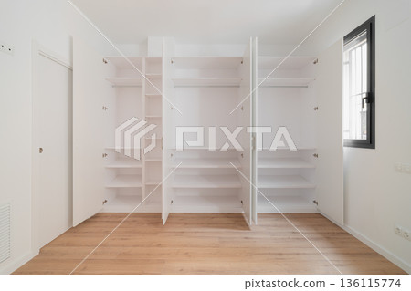 A large white wardrobe with opened doors showcasing interior space and shelves. Example of storage space. Natural light enters through a window. 136115774