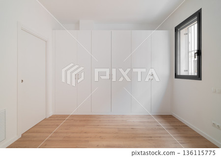 A minimalist interior of a room with white walls and a wooden floor. A large white wardrobe occupies one wall. A window allows natural light to enter. A minimalist interior of a room with white walls and a wooden floor. A large white wardrobe occupies one wall. A window allows natural light to enter. 136115775