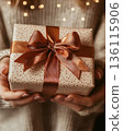 Hands holding beautifully wrapped birthday gift with ribbon. Faceless woman. Surprise, symbolism, festive, joy and emotional birthday celebration, gifting tradition, presents, wishes, holiday concepts 136115906