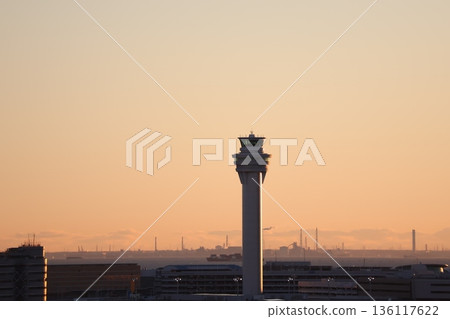 橙色的日出天空、控制塔、機場大樓、大海、船和朦朧的城市景觀 橙色的日出天空、控制塔、機場大樓、大海、船和朦朧的城市景觀 136117622
