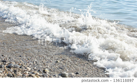 Close-up of a dynamic wave breaking on a pebble beach, capturing the white foam and spray against the clear water. Ideal for themes of nature, travel, or coastal environments. Close-up of a dynamic wave breaking on a pebble beach, capturing the white foam and spray against the clear water. Ideal for themes of nature, travel, or coastal environments. 136118897
