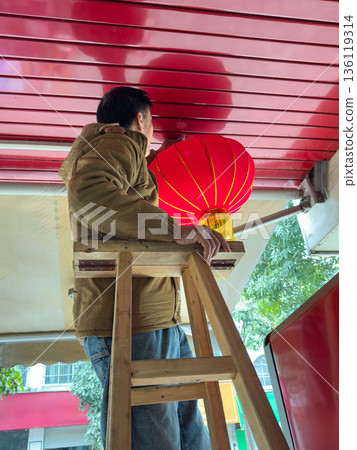 Man Hanging A Vibrant Red Lantern High Up On A Wooden Ladder Man Hanging A Vibrant Red Lantern High Up On A Wooden Ladder 136119314