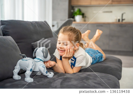 Child interacting with robotic dog while sitting on sofa at home. Artificial intelligence technology and human robot interaction in modern domestic environment. 136119438