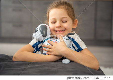 Child hugging robotic dog with affection while sitting on sofa at home. Emotional attachment and digital companionship in modern family life. 136119446